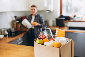 Young redhead woman carrying a delivery box full of fresh organic groceries in the kitchen. A donation box of different products on house background. Express food delivery, online shopping concept