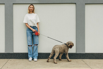 Front view of a young blond woman standing on the sidewalk with her dog on a leash