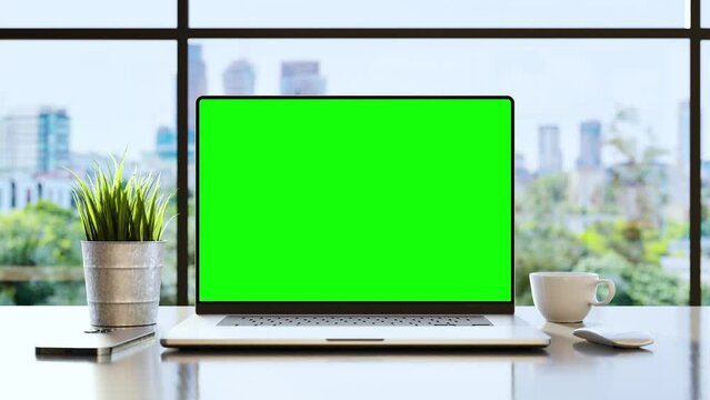 A laptop with an empty green screen on the desk in the office, with trees  swaying in the wind and modern bussines cityscape in the background. Looped video. 3d render