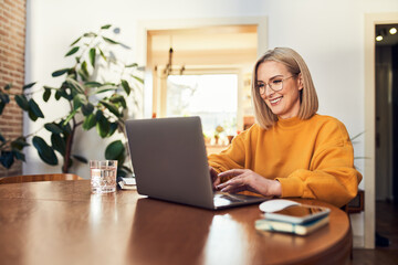 Happy mature woman using laptop while working remotely from home in living room - obrazy, fototapety, plakaty