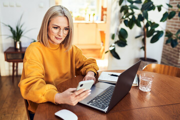 Mature woman using mobile phone while sitting in living room with laptop computer