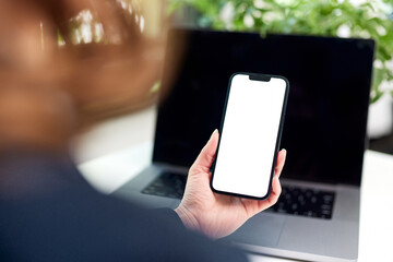 A woman holding a smartphone with a blank white screen in front of the laptop.