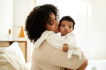 Happy sweet black curly millennial mother with towel kissing little baby, enjoy motherhood