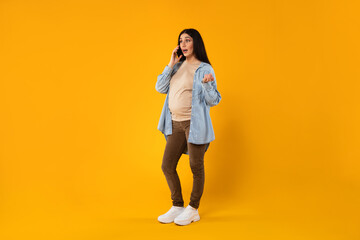 Fototapeta na wymiar Young emotional pregnant woman talking on cellphone and gesturing, yellow studio background, full length, free space