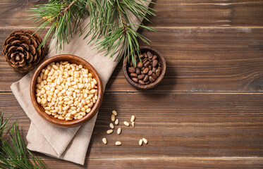 Flat lay of pine nuts in a bowl on napkin  and a handful of unpeeled nuts on a brown wooden background with a branch of pine needles. Healthy diet snack.