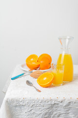 View of plate with cut oranges, bottle and glass with orange juice, juicer, spoon and knife on table with white tablecloth, white background, vertical, with copy space