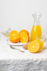 Close-up of table with white tablecloth, bottle and glass with orange juice, juicer, spoon and plate with cut oranges, white background, vertical, with copy space