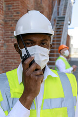 Man standing in reflective uniform and protective mask near unfinished building holding walkie talkie. Portrait of african american man confidently looking at camera outdoors.