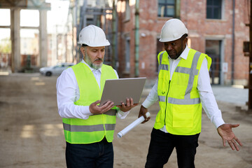 Caucasian senior foreman with wireless laptop discussing about all peculiarities of project with african american male architect at construction area.