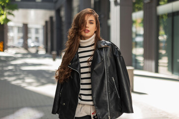 Obraz premium Beautiful young fashionable brunette girl in trendy clothes with a stylish leather jacket and a striped sweater walks in the city on a sunny day