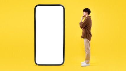 Thoughtful Guy Looking At Big Smartphone Screen On Yellow Background
