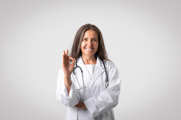 Happy senior woman therapist in white coat with stethoscope showing ok sign, posing isolated on gray background