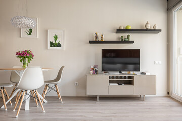 Cozy living room with panoramic windows, a dining table and chairs against the backdrop of a wall with a TV, shelves and decorative accessories and flowers. Concept of a stylish apartment interior