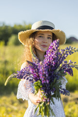 Beautiful young girl in a white dress, straw hat, with picnic and bouquet of purple wild flowers on a meadow. Summertime, golden hour, sunset