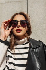 Sunny urban female portrait of a beautiful fashionable woman in stylish clothes with a sweater and leather jacket puts on fashion sunglasses