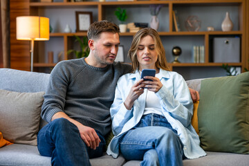 Happy young couple sitting on a couch at home, using mobile phone