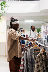 African american man searching for trendy outfit in clothing store. Customer browsing through shirts hanging on rack to choose casual apparel in shopping mall fashion boutique