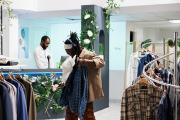 Cheerful buyer checking out shirt while listening to music in headphones in clothing store. African...