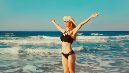 Summer vacation, happy smiling woman in bikini swimsuit and straw hat raising her hands up on the beach on sea coast background on sunny day
