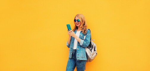 Portrait of stylish modern smiling young woman with smartphone wearing jean jacket on yellow background