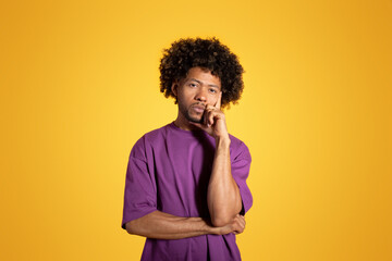 Fototapeta na wymiar Pensive bored sad mature black curly man in purple t-shirt thinks, chooses isolated on yellow background
