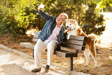 Glad european old man in casual enjoys walking with dog, sit on bench in park, taking photo on smartphone