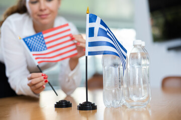 Unrecognizable unfocused woman preparing room for international negotiations and communication discussions of leaders. Lady sets miniatures flags of Greece and USA on table.