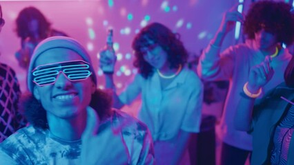 Medium close-up shot of young biracial male student in neon glasses dancing energetically at student rave party, diverse friends dancing with beer, DJ playing, in pink and blue disco ball lights