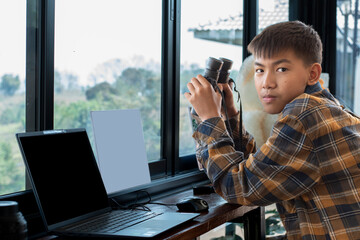 Asian cute boy in plaid shirt holds binoculars, sitting near glass window in living room, ready to see birds on trees outside his house, concept for relaxing and recreational activity.
