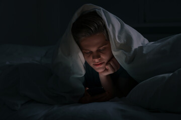 Teenage boy using smartphone under blanket on bed at night. Internet addiction