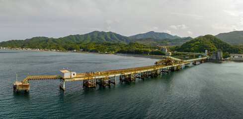 Long loading pier with rusted and equipment stretches into sea on coast