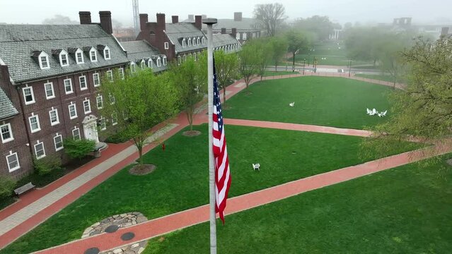 Rotational aerial shot of stagnant American flag on academic lawn of foggy university. Walkable college campus with green spaces and historic brick dorms. University of Delaware in Newark, Delaware.