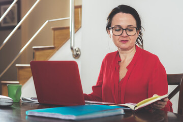 Woman in red dress with red laptop and notebook comparing data at home. Business concept.