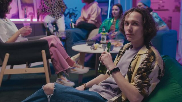 Medium portrait shot of young Caucasian man with curly hair, in ripped jeans, bright shirt sitting on beanbag at house party, drinking beer, then looking at camera, and friends chatting in background