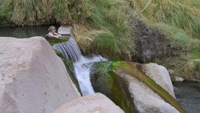 Girl Relaxing by Huge Rocks, Grassand Waterfall in the Puritama Termas Hot Springs, Northern Chile.