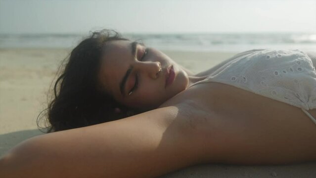 Close-up shot of a young beautiful Indian girl lying on the sand on a beach and opening her eyes. Indian Ocean in the background a suny day. 