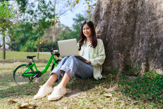 Women sitting under tree to working and surfing social media on laptop after cycling in the park