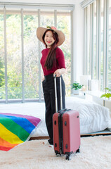 Asian young happy cheerful female in casual travel outfit standing preparing holding trolley luggage together going on vacation on holiday weekend with rainbow pride LGBTQ freedom flag