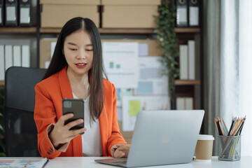 Asian woman talking mobile phone and working on laptop and calculator on table in office.