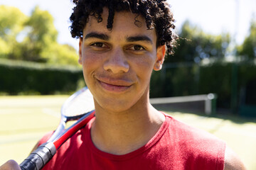 Close-up portrait of confident biracial young man with tennis racket smiling in tennis court