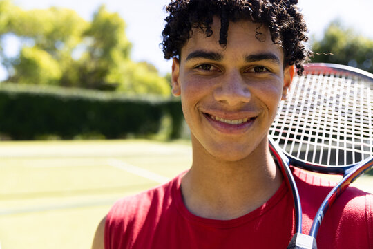 Close-up of biracial young man with tennis racket smiling in tennis court and looking at camera