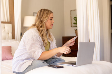 Caucasian plus size young woman doing online shopping with credit card and laptop on bed