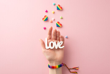 A first person top view of a woman's hand wearing a rainbow bracelet and holding the word "love" on her palm and accessories against a pastel pink background symbolizing support for the LGBT community