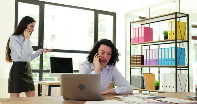 Young businessman is talking on phone while sitting at table and working on laptop in coworking space with colleagues. Manager answering call in shared office with colleagues