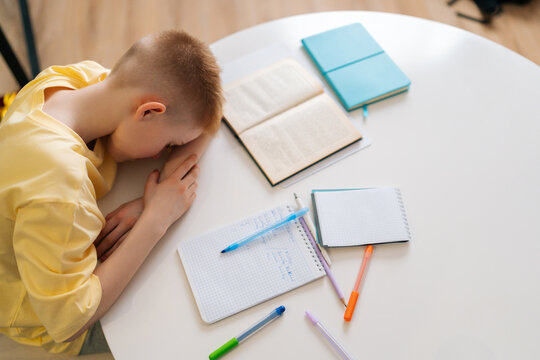 High-angle view of tired student pupil boy lying on desk filled with books training material. Frustrated little child boy sleeping while doing homework with head on table. Fatigue, education concept
