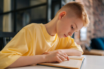 Side view face of adorable pupil schoolboy reading paper book following lines with finger along...