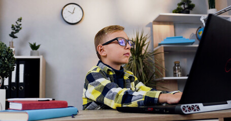 Portrait of attractive teen smart boy in eyeglasses which sitting at his workplace and doing homework using computer