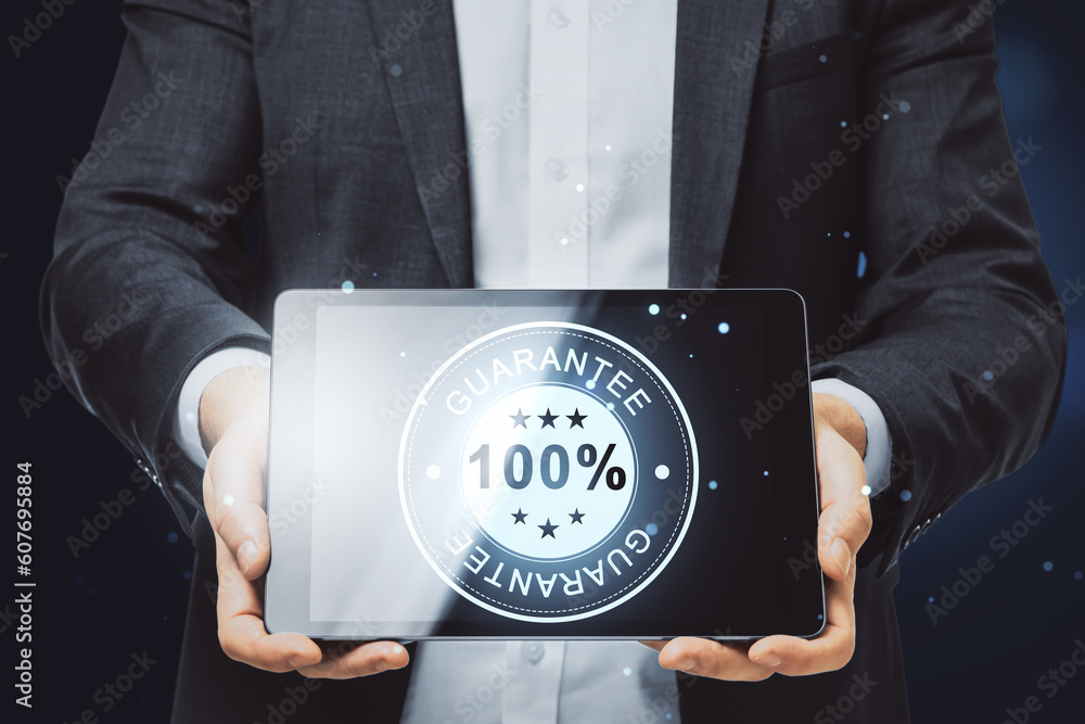 Poster close up of businessman hand holding tablet with glowing 100% guarantee stamp hologram on dark backg