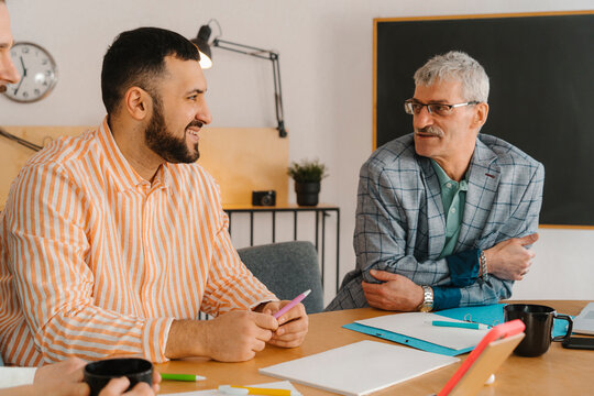 Male colleagues laugh and talk sitting at studio table