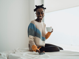 Smiling woman holding coffee cup and using smartphone on bed